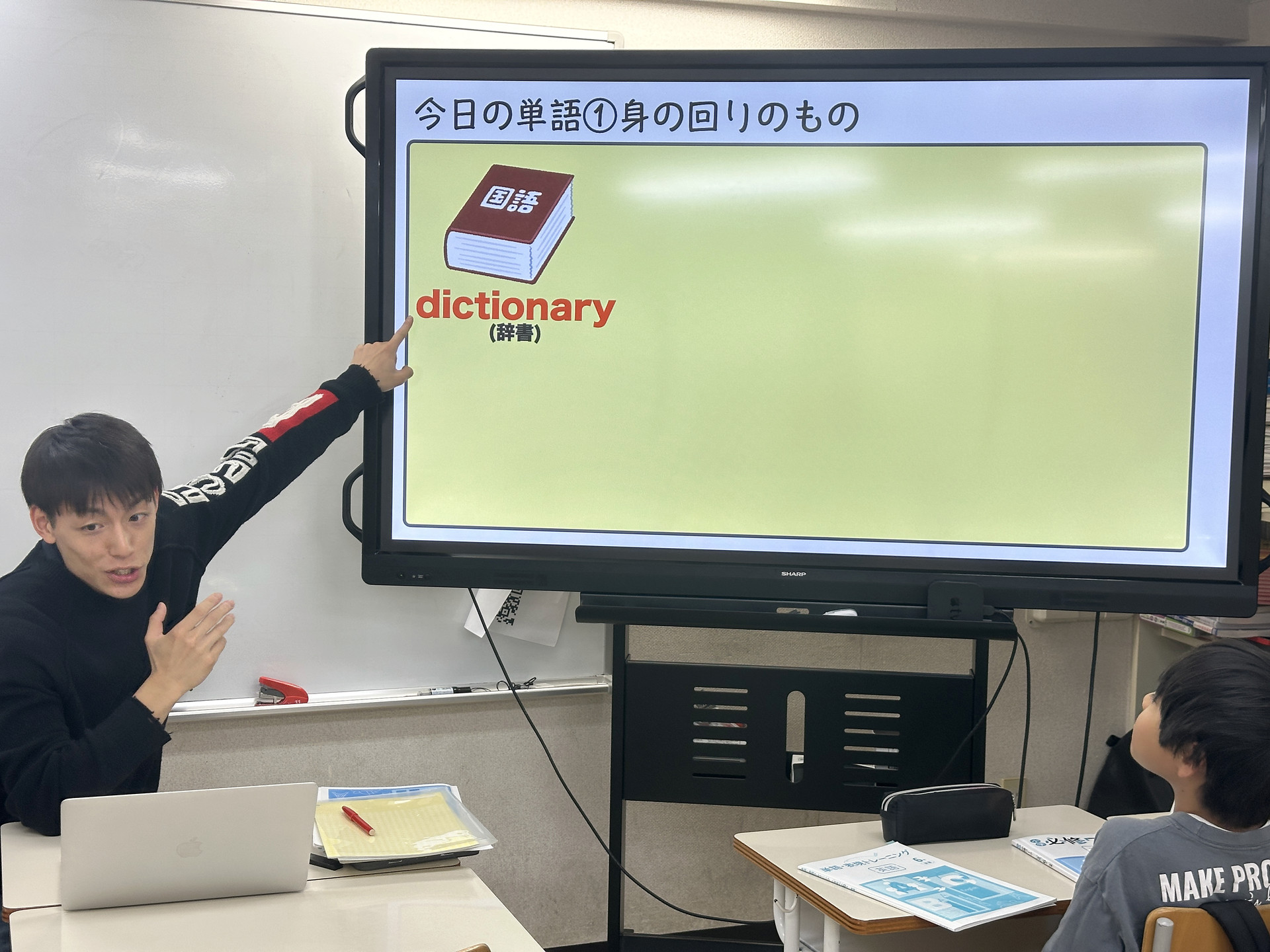 画面の単語を指し示す先生と、授業に参加する小学生。